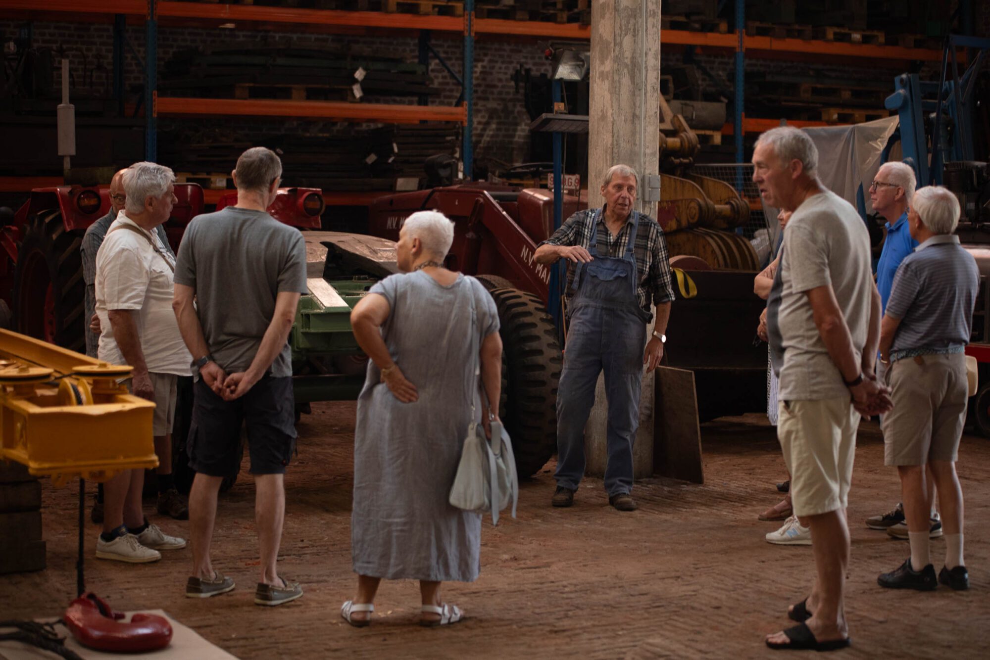 Group of visitors listening to a volunteer guide among historic port vehicles and machinery inside a warehouse.