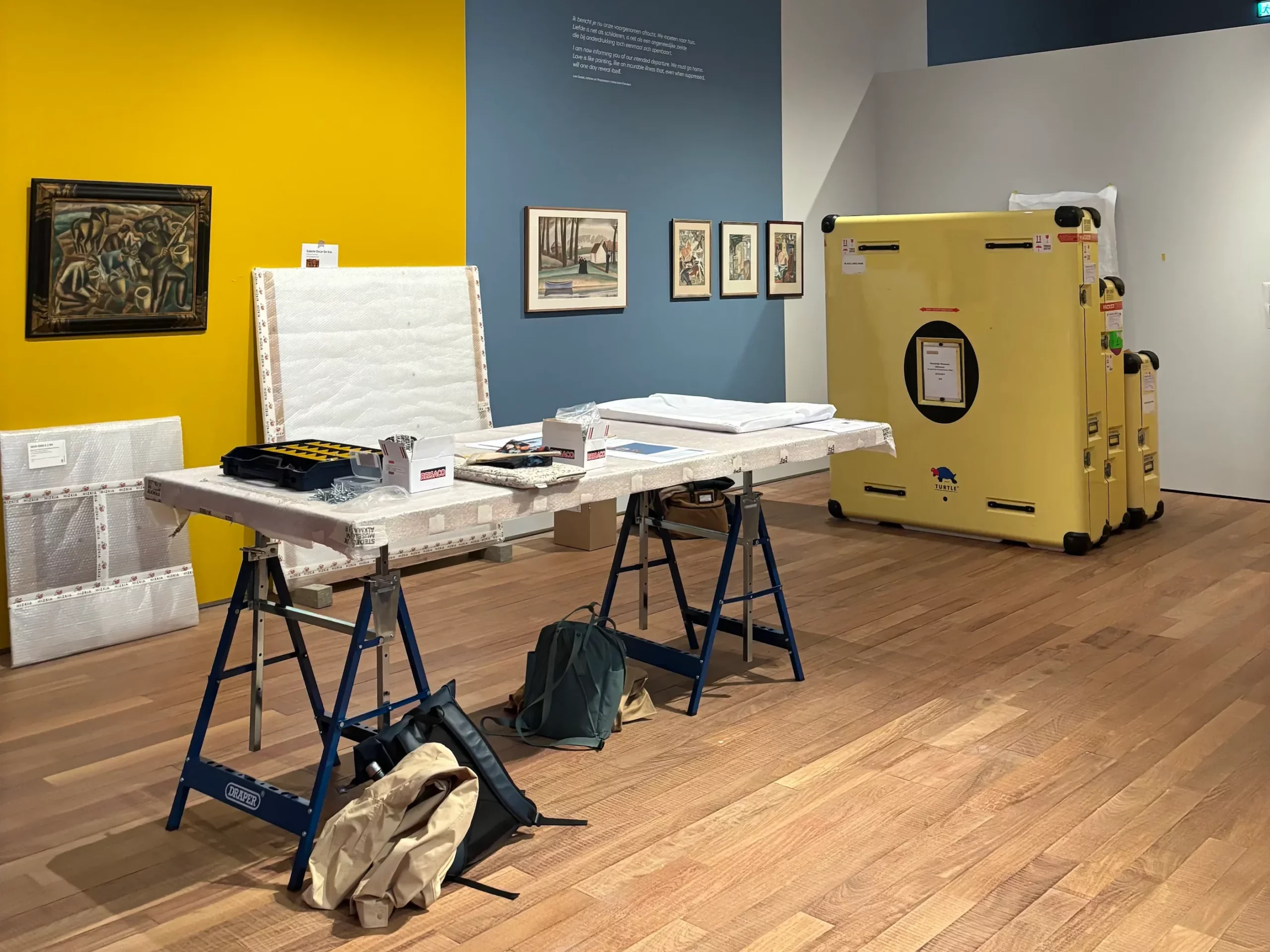 Zaalopbouw van een tentoonstelling met ingepakte kunstwerken, transportkisten en restauratiemateriaal tijdens de installatie van schilderijen in een museumruimte.